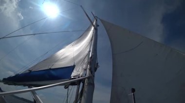 Yatlar rüzgarda dalgalanır, parlak mavi bir gökyüzüne ve parlayan güneşe karşı batar. Dinamik sahne, lüks, macera.