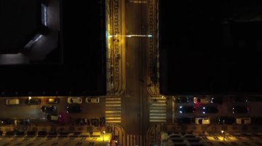 Bir İHA gece bir şehir caddesinin üzerinde uçuyor, çatılar ve kavşaklar üzerinde, parlayan sokak lambaları ve aşağıdaki ara sıra araba hareketleri, kentsel gece hayatı atmosferini gösteriyor.