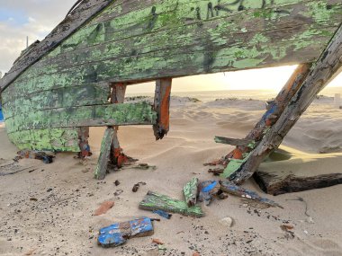 Eski terk edilmiş ahşap bir tekne, kısmen kumsalda gömülü. Soluk yeşil boya soyuluyor, çatlamış ahşabı ortaya çıkarıyor. Güneş ışığı kumun dokusunu vurgular, enkaz parçalarını..