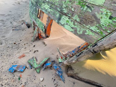Eski terk edilmiş ahşap bir tekne, kısmen kumsalda gömülü. Soluk yeşil boya soyuluyor, çatlamış ahşabı ortaya çıkarıyor. Güneş ışığı kumun dokusunu vurgular, enkaz parçalarını..