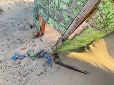 Eski terk edilmiş ahşap bir tekne, kısmen kumsalda gömülü. Soluk yeşil boya soyuluyor, çatlamış ahşabı ortaya çıkarıyor. Güneş ışığı kumun dokusunu vurgular, enkaz parçalarını..