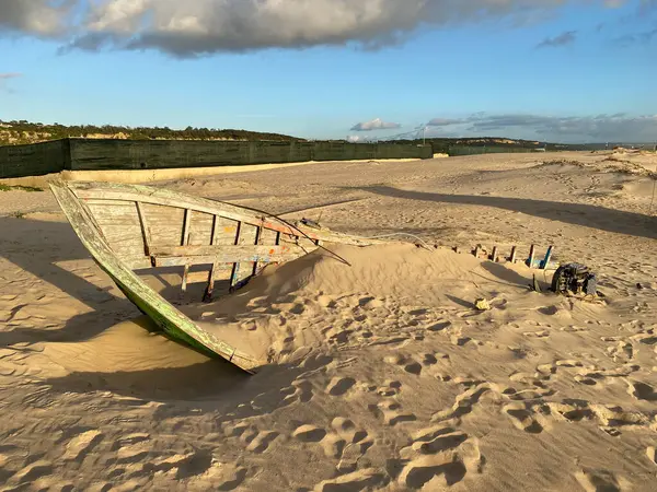 Eski terk edilmiş ahşap bir tekne, kısmen kumsalda gömülü. Soluk yeşil boya soyuluyor, çatlamış ahşabı ortaya çıkarıyor. Güneş ışığı kumun dokusunu vurgular, enkaz parçalarını..
