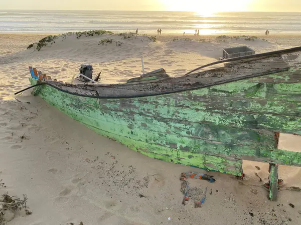 Eski terk edilmiş ahşap bir tekne, kısmen kumsalda gömülü. Soluk yeşil boya soyuluyor, çatlamış ahşabı ortaya çıkarıyor. Güneş ışığı kumun dokusunu vurgular, enkaz parçalarını..
