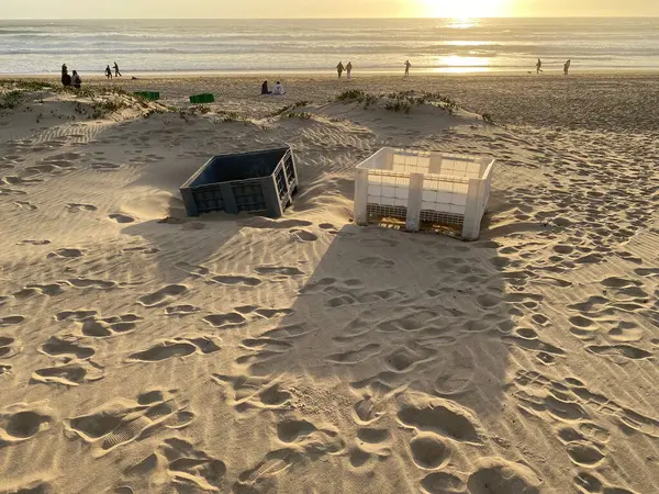 Terk edilmiş iki plastik sandık kumsalda, sahilde, gölgelerde, gün batımının altın ışığında kısmen gömülü duruyor. Ayak izleri kum tepelerini kaplıyor..