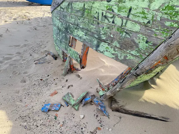 Eski terk edilmiş ahşap bir tekne, kısmen kumsalda gömülü. Soluk yeşil boya soyuluyor, çatlamış ahşabı ortaya çıkarıyor. Güneş ışığı kumun dokusunu vurgular, enkaz parçalarını..