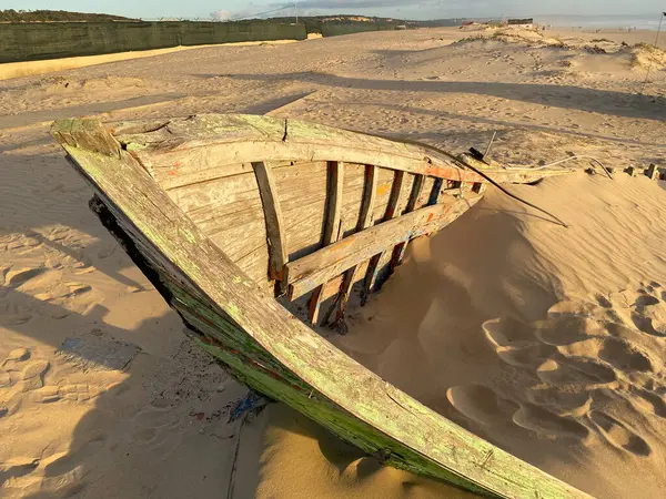 Eski terk edilmiş ahşap bir tekne, kısmen kumsalda gömülü. Soluk yeşil boya soyuluyor, çatlamış ahşabı ortaya çıkarıyor. Güneş ışığı kumun dokusunu vurgular, enkaz parçalarını..