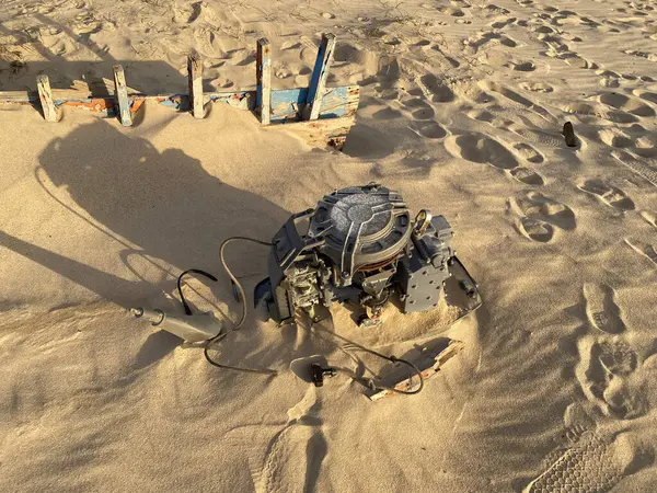 Paslı sökülmüş bir tekne motoru kumsaldaki kumda kısmen yatıyor. Terk edilmiş bir teknenin ahşap yapısı sıcak güneş ışığıyla yıkanır..