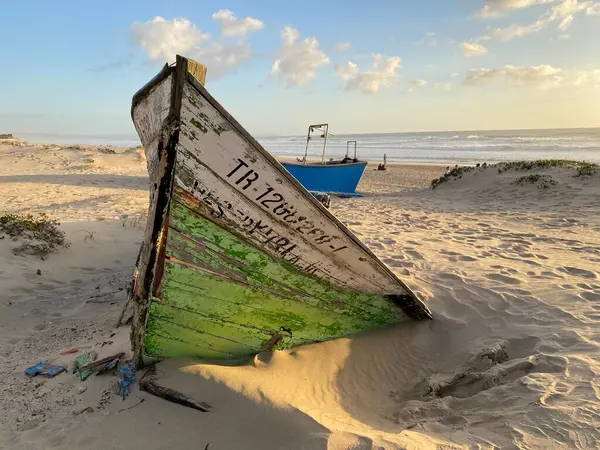 Eski terk edilmiş ahşap bir tekne, kısmen kumsalda gömülü. Soluk yeşil boya soyuluyor, çatlamış ahşabı ortaya çıkarıyor. Güneş ışığı kumun dokusunu vurgular, enkaz parçalarını..