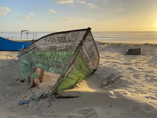 Eski terk edilmiş ahşap bir tekne, kısmen kumsalda gömülü. Soluk yeşil boya soyuluyor, çatlamış ahşabı ortaya çıkarıyor. Güneş ışığı kumun dokusunu vurgular, enkaz parçalarını..