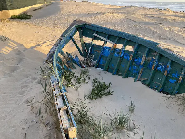 Eski terk edilmiş ahşap bir tekne, kısmen kumsalda gömülü. Soluk yeşil boya soyuluyor, çatlamış ahşabı ortaya çıkarıyor. Güneş ışığı kumun dokusunu vurgular, enkaz parçalarını..