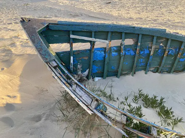 Eski terk edilmiş ahşap bir tekne, kısmen kumsalda gömülü. Soluk yeşil boya soyuluyor, çatlamış ahşabı ortaya çıkarıyor. Güneş ışığı kumun dokusunu vurgular, enkaz parçalarını..
