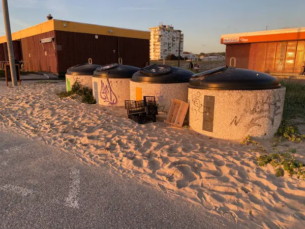 Batan güneşle aydınlatılan bir kumsal yakınındaki kumlu zeminde grafiti ile çöp kutularını ayıran bir sıra. Atılmış sandıklar ve yakınlarda bir palet vardı. Costa da Caparica, Portekiz. 16.2.2025 