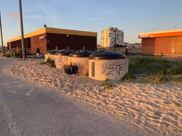Batan güneşle aydınlatılan bir kumsal yakınındaki kumlu zeminde grafiti ile çöp kutularını ayıran bir sıra. Atılmış sandıklar ve yakınlarda bir palet vardı. Costa da Caparica, Portekiz. 16.2.2025 