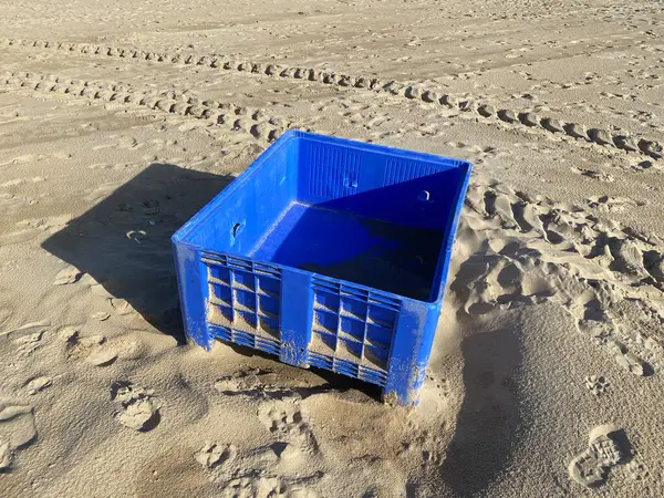 Terk edilmiş mavi balık plastik sandığı kumsalda. Gölgeler, güneş. Çekici tekerlek izleri, kumulları kaplayan ayak izleri..