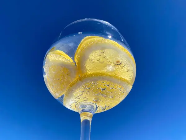 Parlak mavi gökyüzüne karşı limon dilimli, köpüklü narenciye içeceği yakın plan. Meyveye yapışan küçük kabarcıklar taze ve ferahlatıcı hissi arttırır. Yaz, su, sağlıklı içecek.
