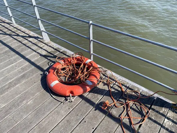Ahşap iskelede aşınmış halatla yıpranmış bir can simidi. Güneş ışığı ve gölgeler ham, denizsel bir atmosfer yaratır. Kurtarma, ihmal ya da zaman akışının kavramsal görüntüsü.