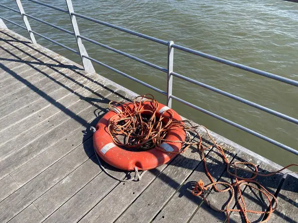 Ahşap iskelede aşınmış halatla yıpranmış bir can simidi. Güneş ışığı ve gölgeler ham, denizsel bir atmosfer yaratır. Kurtarma, ihmal ya da zaman akışının kavramsal görüntüsü.