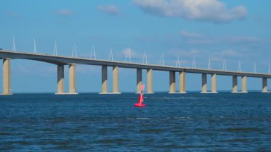 Vasco da Gama Köprüsü nehrin üzerinde yüzerek soyut bir mimari arkaplan oluşturur. Temiz yatay çizgiler, derin mavi su ve minimalist dizayn gökyüzü ve renkli şamandıralarla harmanlanmış..