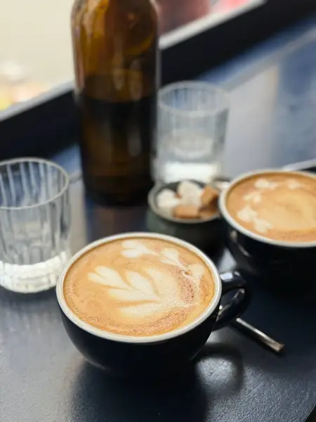 Sıcak bir fincan latte, koyu renkli bir masanın üzerinde narin köpük sanatı, cam süsler ve şeker küpleriyle çevrili. Yumuşak sabah ışığı, sakin ve rahat bir kahvaltı atmosferini vurgular.