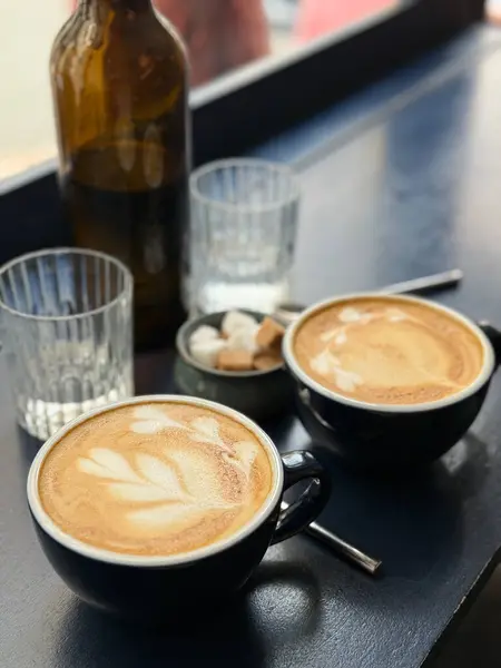 Sıcak bir fincan latte, koyu renkli bir masanın üzerinde narin köpük sanatı, cam süsler ve şeker küpleriyle çevrili. Yumuşak sabah ışığı, sakin ve rahat bir kahvaltı atmosferini vurgular.