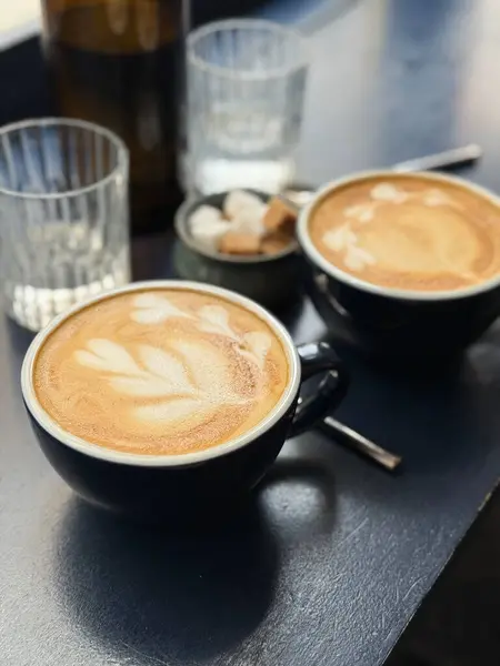 Sıcak bir fincan latte, koyu renkli bir masanın üzerinde narin köpük sanatı, cam süsler ve şeker küpleriyle çevrili. Yumuşak sabah ışığı, sakin ve rahat bir kahvaltı atmosferini vurgular.