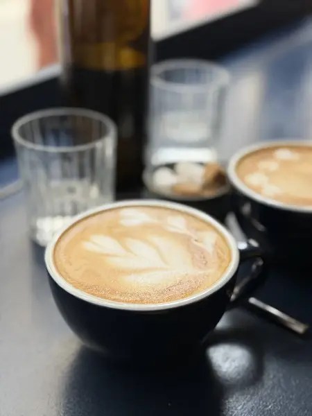 Sıcak bir fincan latte, koyu renkli bir masanın üzerinde narin köpük sanatı, cam süsler ve şeker küpleriyle çevrili. Yumuşak sabah ışığı, sakin ve rahat bir kahvaltı atmosferini vurgular.