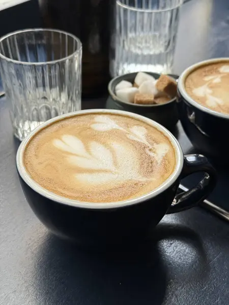 Sıcak bir fincan latte, koyu renkli bir masanın üzerinde narin köpük sanatı, cam süsler ve şeker küpleriyle çevrili. Yumuşak sabah ışığı, sakin ve rahat bir kahvaltı atmosferini vurgular.