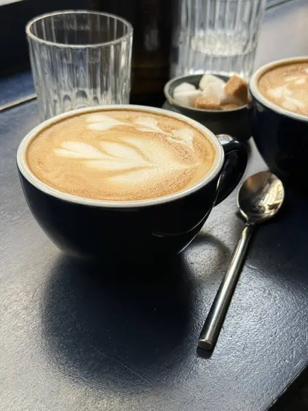 Sıcak bir fincan latte, koyu renkli bir masanın üzerinde narin köpük sanatı, cam süsler ve şeker küpleriyle çevrili. Yumuşak sabah ışığı, sakin ve rahat bir kahvaltı atmosferini vurgular.