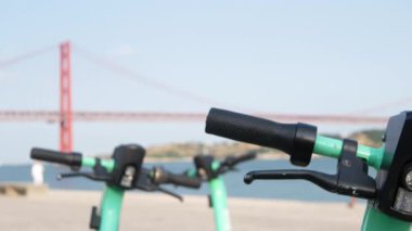 Yakın plan elektrikli scooter gidonu, akıllı şehir hareket kabiliyeti, modern gidip gelme, çağdaş şehir yaşamında ekolojik bilinçli ulaşım. Arka planda 25 de Abril Köprüsü, Lizbon, Portekiz.