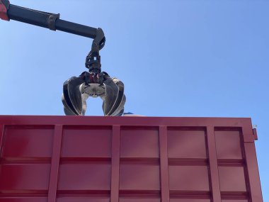 Hidrolik endüstriyel pençeler, açık mavi gökyüzüne karşı kırmızı bir konteynırın üzerinden metal parçaları kapıyor. Atık yönetimi, geri dönüşüm veya inşaat işleri için ağır makineler kullanılıyor.