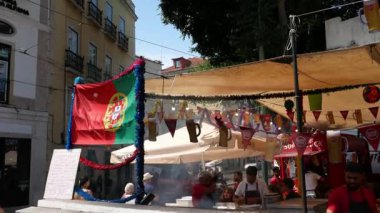Lizbon, Portekiz 13 Haziran 2025: Alfama 'daki sokak restoranı, Portekiz bayrakları ve şenlikli çelenklerle süslenmiş. İnsanlar eski şehrin göbeğinde güneş gözlüğü altında yemek tezgahlarından hoşlanırlar..
