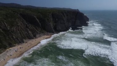 Portekiz 'in Sintra kentindeki Grande ve Adraga Sahili' ndeki Atlantik Okyanusu 'nun yüksek kayalık kıyılarının insansız hava aracı panoraması. İnsanlar sahilde dinleniyor. Siyah kayaların altındaki sarı kumların üzerinde uzun beyaz dalgalar. Hava görünümü.