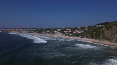 Okyanus kıyı şeridi üzerindeki drone manzarası, bir yaz plajı tatil köyüne yaklaşıyor. Yumuşak dalgalar kumlu sahile doğru akıyor, insanların evlerle benekli yeşil bir yamacın yakınında güneşin tadını çıkardıkları yere..