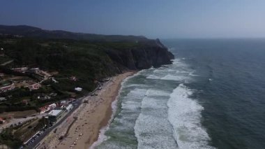 Portekiz 'in Sintra kentindeki Grande ve Adraga Sahili' ndeki Atlantik Okyanusu 'nun yüksek kayalık kıyılarının insansız hava aracı panoraması. İnsanlar sahilde dinleniyor. Siyah kayaların altındaki sarı kumların üzerinde uzun beyaz dalgalar. Hava görünümü.