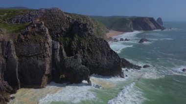 Portekiz 'in Sintra kentindeki Grande ve Adraga Sahili' ndeki Atlantik Okyanusu 'nun yüksek kayalık kıyılarının insansız hava aracı panoraması. İnsanlar sahilde dinleniyor. Siyah kayaların altındaki sarı kumların üzerinde uzun beyaz dalgalar. Hava görünümü.