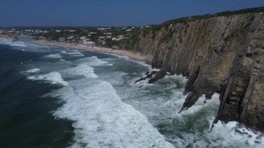 Portekiz 'in Sintra kentindeki Grande ve Adraga Sahili' ndeki Atlantik Okyanusu 'nun yüksek kayalık kıyılarının insansız hava aracı panoraması. İnsanlar sahilde dinleniyor. Siyah kayaların altındaki sarı kumların üzerinde uzun beyaz dalgalar. Hava görünümü.