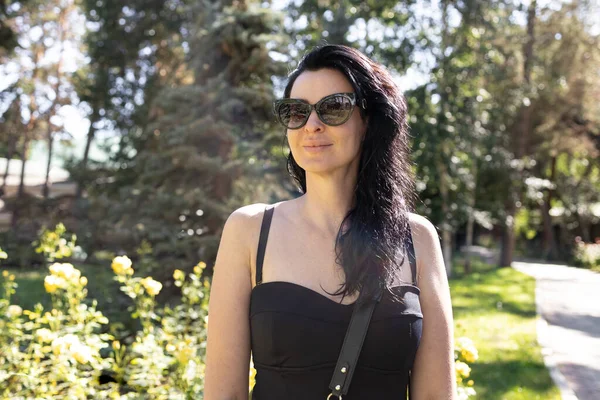 Güzel, uzun saçlı esmer, kayışlı ve güneş gözlüklü siyah bir elbise giymiş. Parkta güneşli havanın keyfini çıkar. Kendine güvenen çekici bir kadın tatil köyünde yürüyor. Yaz tatili konsepti.