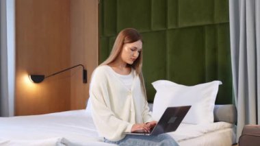 Uzun saçlı genç bayan, yeşil ve ahşap dekorlu, modern, rahat bir otel odasında dizüstü bilgisayarının üzerinde oturuyor..