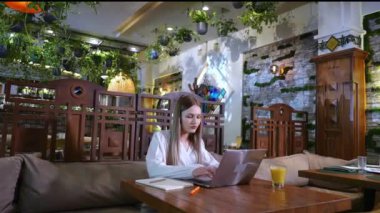 Genç bir kadın tüccar, lüks bir restoranda, ahşap mobilyaları, bitkileri ve yeşillikleri olan kahvaltısını beklerken dizüstü bilgisayarına odaklanıyor.