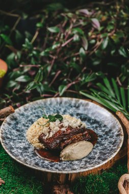 Yeşilliklerle servis edilen bol pişmiş pirzola, ptitim ve kremalı ezme, bereketli yeşilliklerle çevrili, mavi ve beyaz desenli bir tabakta servis edilir.