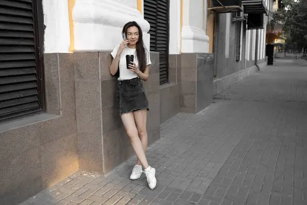 Çekici Asyalı kız kot etek ve beyaz tişört giyiyor. Elinde cep telefonu var ve caddede kahve içiyor..