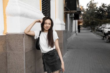 Beyaz gömlekli ve siyah kotlu genç bir kadın şehir caddesinde duvara yaslanıyor, eli başının üzerinde dinleniyor.