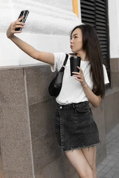 Beyaz tişört ve siyah etek giyen uzun saçlı bir kadın şehirde kahve fincanı tutarken selfie çekiyor.