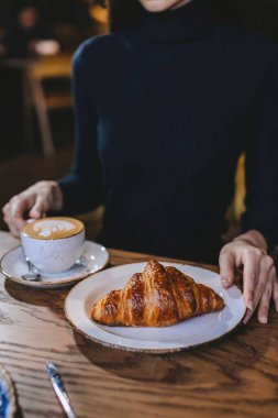 Siyah giyinmiş, altın kruvasan ve sıcak bir restoranda sıcak bir fincan kahvenin tadını çıkaran, serin bir gün için mükemmel bir ortam ile çevrili isimsiz bir kişi..