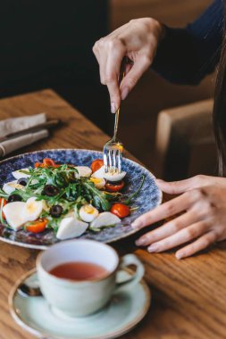 Bir el yeşillik, kiraz domatesleri ve yumurtalarla dolu güzel bir tabaktan bir parça salata seçiyor, yanında da sıcak bir kafe masasında bir fincan çay..