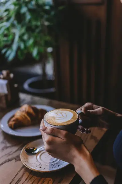 Bir kişi ahşap bir masada oturuyor, köpük sanatı ile karışık bir latte tutuyor. Yakınlardaki bir tabakta taze pişmiş kruvasan duruyor, rahat bir kafe ortamında yeşilliklerle çevrili..