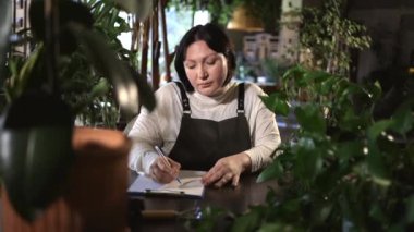 Akıllı bahçıvan ahşap masada notlar almaya odaklanır. Etrafı çeşitli kapalı bitkilerle çevrilidir. Küçük işletme sahibi işten sonra stokları kontrol ediyor. Müteşebbis, ertesi gün için program hazırlıyor.