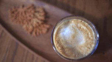 Bilinmeyen kişi kahve bardağında karıştırıyor. Havadar kremalı ve ahşap tepside gevrek ikram edilen latte camının en üst görüntüsü. Konforlu kafe kavramı.