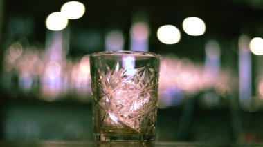Kristal eski moda cam parlak ahşap masaya oturur, canlı bar atmosferindeki sıcak ışığı yansıtır. Arka planda bulanık ışıklar var, canlı ayarları güçlendiriyor. Buzlu viski..