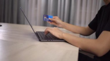 Genç bir kadın masa başında oturmuş, bilgisayarını kullanarak online işlemi tamamlıyor. Klavyeye yazarken kredi kartını tutan kadının elini kapat ve işine odaklan. Çevrimiçi satın alma kavramı.